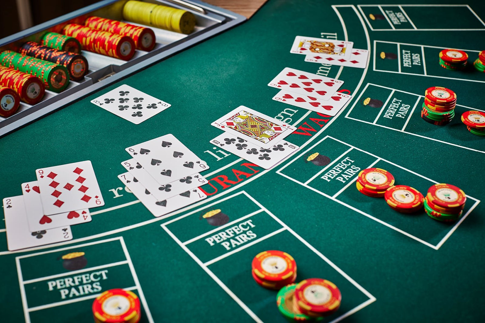 A close-up photo of a green blackjack casino table showing several hands of playing cards, colorful poker chips stacked in betting areas labeled "Perfect Pairs," and a chip tray filled with red, yellow, and green chips—depicting an active game setting.